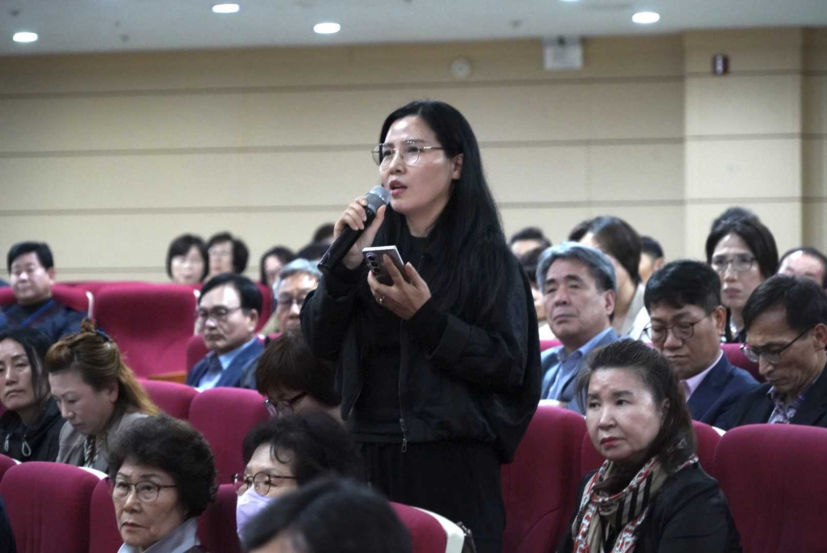 객석의 한 참석자가 휴대전화를 손에 든 채 마이크로 이야기하고 있다.